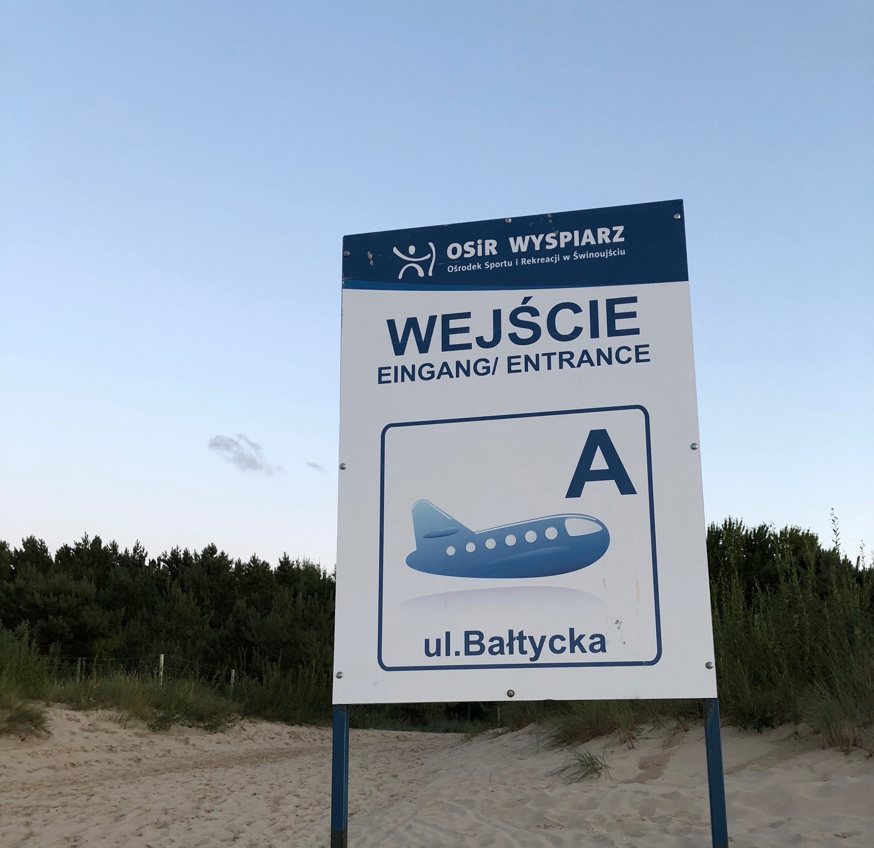 Tablica oznaczająca wejście na plażę z napisem: Wejście, Eingang, Entrance, A, ul. Bałtycka, i wizarunkiem samolotu. W tle fragment piaszczystej wydmy porośniętej roślinnością.